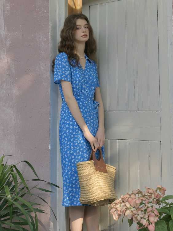 Model standing calmly in front of a wooden door, wearing a blue floral belted dress and holding a straw tote bag. Soft shadows and pastel tones create a serene, fairytale-inspired atmosphere.