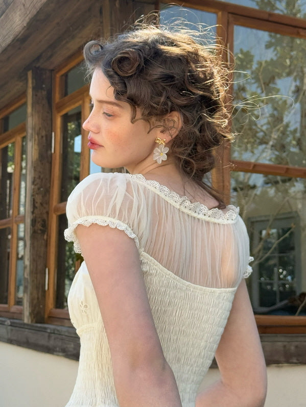 Back view of a princesscore ivory dress for women featuring sheer puff sleeves, delicate lace edging, and smocked bodice, captured in a soft vintage-inspired cottage setting.