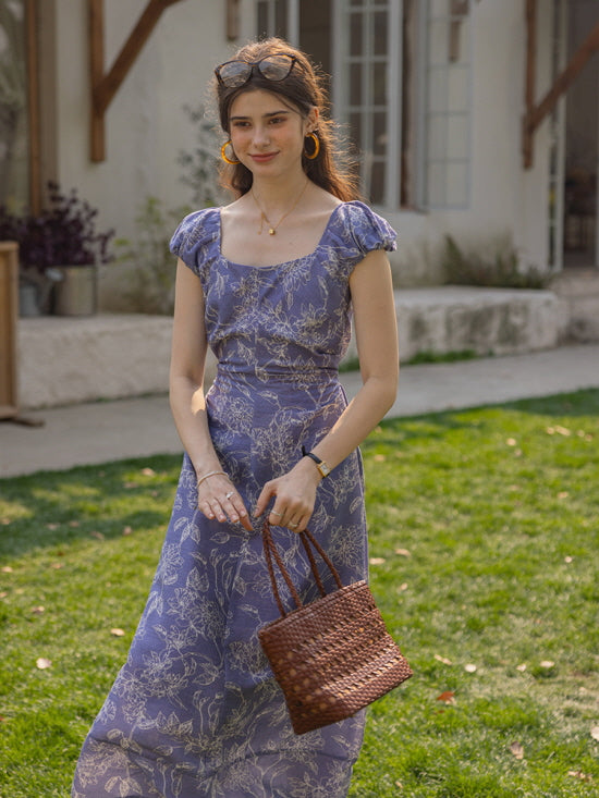 Woman in a blue floral puff-sleeve dress holding a woven bag, standing in a sunlit garden with a soft, cottagecore aesthetic and a charming countryside ambiance.