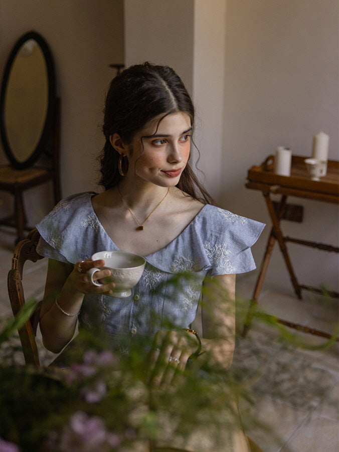 A young woman with soft, wavy hair sips delicately from a ceramic teacup, her gaze wandering thoughtfully. She wears a light blue floral cotton dress with ruffled sleeves, seated in a vintage-inspired room adorned with warm, wooden furniture and gentle, natural light filtering through the plants in the foreground