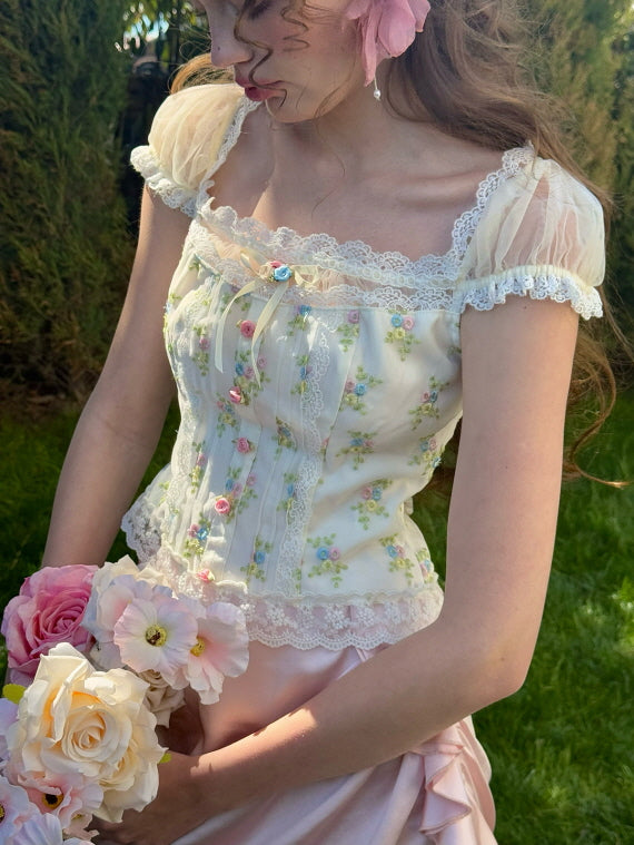 A close-up of a floral corset top adorned with lace detailing and pastel floral embroidery, styled with a pink ruched satin skirt and complemented by a bouquet of roses, evoking a romantic, fairytale aesthetic.