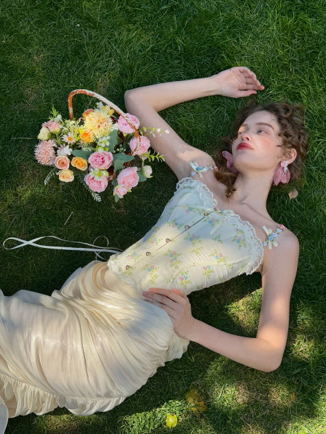 Romantic cream ruched satin skirt styled with a floral embroidered corset top, lying on grass beside a basket of pastel flowers – fairytale garden outfit for women