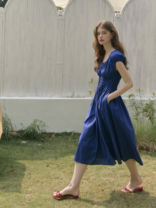 Model wearing a shirred navy midi dress with pockets, walking through a sunlit garden with gentle movement. Elegant fairytale-inspired look with a natural, carefree mood.