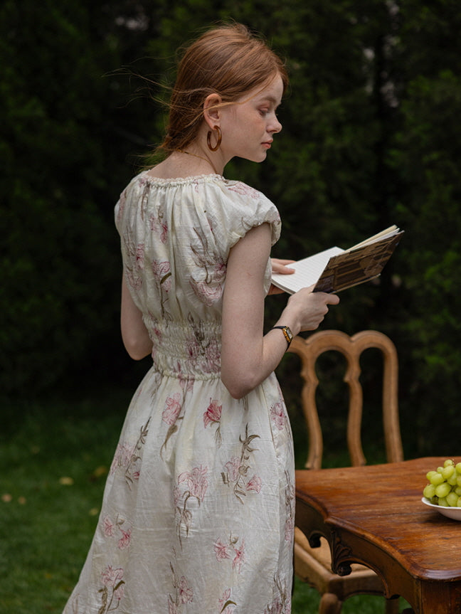 Back view of a woman reading a book while wearing a floral cottagecore dress with cap sleeves and a smocked waist. Dreamy, nature-inspired setting.