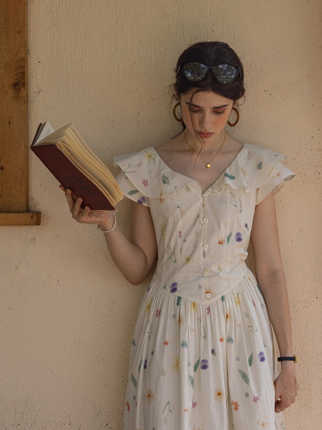 Young woman leaning against a rustic wall, holding a book while wearing a vintage-inspired floral dress with flutter sleeves, capturing a serene and thoughtful mood.