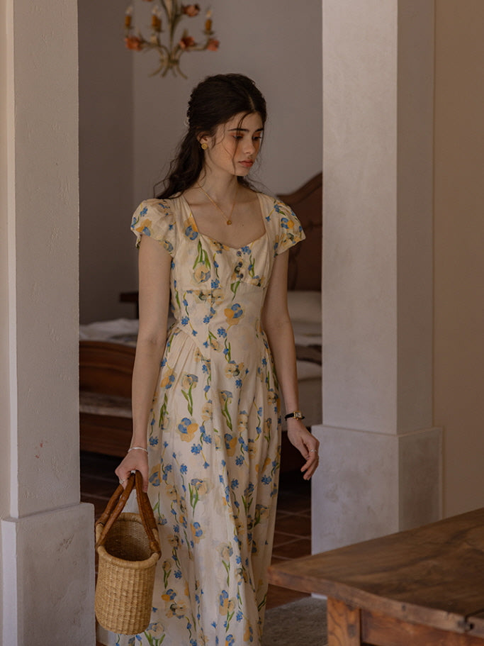 The model walks gracefully through a rustic bedroom, holding a woven basket bag. Her cream floral dress with blue and yellow flower prints flows gently, capturing a serene, cottagecore vibe with warm, natural tones and soft, afternoon light.