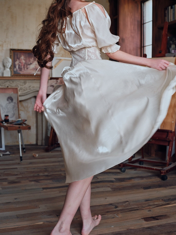 A barefoot woman twirling in an ivory satin dress with a lace corset, her hair flowing as the skirt sways gracefully in a vintage art studio setting.
