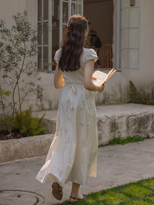 Back view of a woman walking toward a rustic house, wearing a white cotton dress with lace embroidery details and a tied ribbon at the back, holding an open book while strolling through a grassy garden under soft afternoon light.