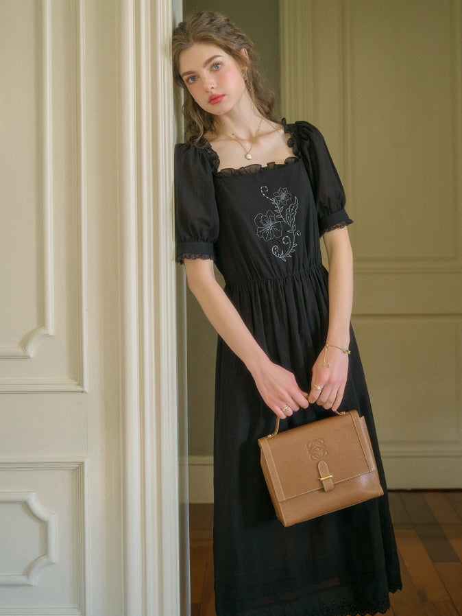 A young woman with wavy hair wearing a black puff sleeve midi dress with white floral embroidery on the bodice, standing indoors against a white wall, holding a brown vintage-style handbag.