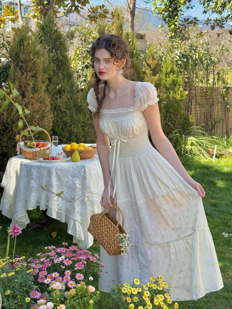A woman wearing a romantic cottagecore dress in ivory with lace trim and embroidered flowers, standing beside a vintage picnic table in a fairytale garden filled with spring blooms.