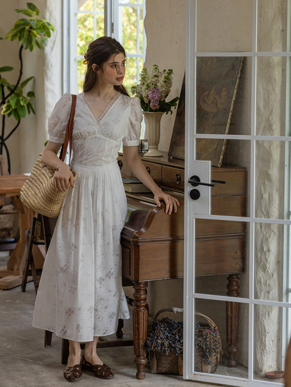 A woman standing by a wooden piano while wearing a white cotton dress adorned with delicate floral embroidery, carrying a woven straw bag and gazing thoughtfully out the window in a cottage-style room.