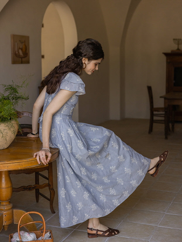 The model playfully lifts her foot while seated on a wooden table, her floral embroidered dress cascading softly around her. The light blue dress with a ruffled neckline and delicate floral patterns evokes a whimsical, fairytale-like mood in the rustic room setting.