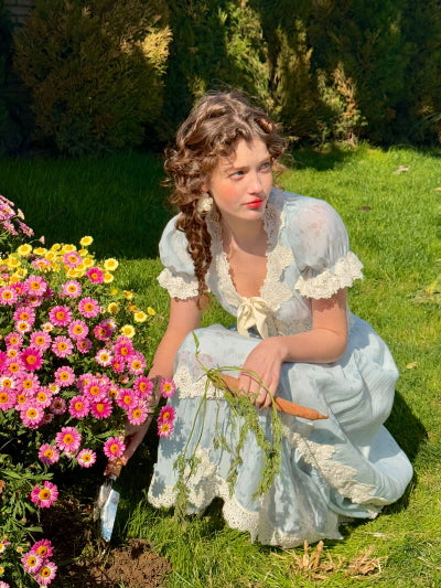 A woman in a romantic blue lace-trimmed dress sitting on green grass with a wooden bowl of colorful cherry tomatoes and carrots nearby, styled in a dreamy countryside aesthetic.
