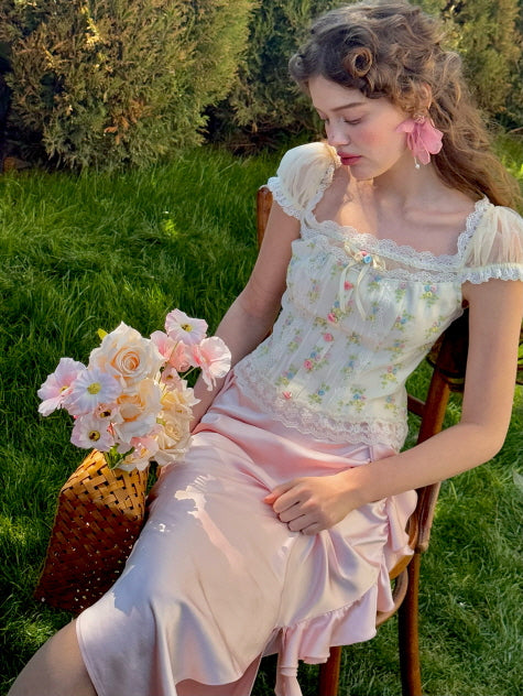 A model seated in a garden chair, wearing a floral lace corset top with sheer puff sleeves and a pink ruched satin skirt, holding a bouquet of pastel roses, evoking a soft, romantic aesthetic.