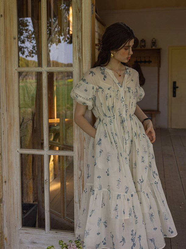 Woman in a cream floral print dress with puff sleeves and tiered skirt, gently touching the fabric while standing in front of open French doors. The scene exudes a nostalgic, countryside cottagecore aesthetic.