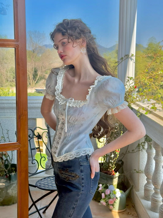 A model poses in a pastel blue corset top with lace accents and puff sleeves, styled with dark denim jeans featuring floral embroidery, creating a dreamy, vintage-inspired look on a sunlit balcony.