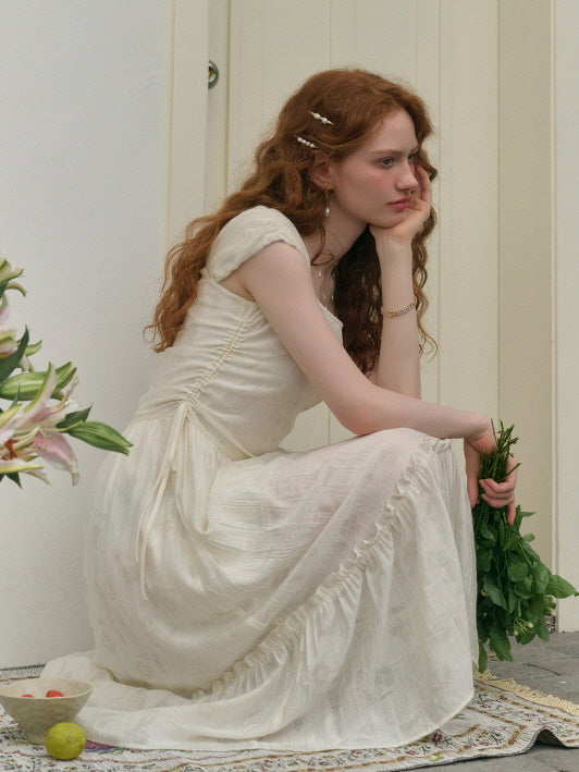 A young woman with long curly red hair sits thoughtfully on a rug, wearing a soft cream-colored romantic dress with ruffle details and holding fresh greenery, against a light backdrop.