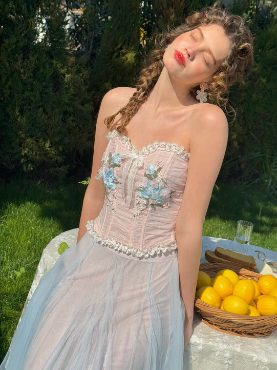 Romantic woman in a pink corset dress with blue floral embroidery and soft tulle skirt, sitting by a table with lemons in sunlight – vintage spring garden fashion