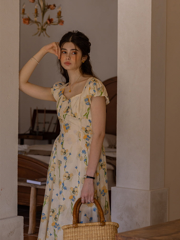 The model leans casually against a column, her expression serene and reflective. She holds a woven basket bag while wearing a cream floral dress with blue and yellow flowers, capturing a dreamy, fairytale-like ambiance amidst a rustic bedroom setting.