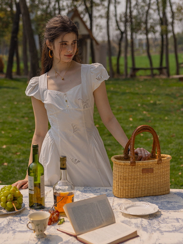 A woman in a vintage-inspired white embroidered dress with puff sleeves, standing at an outdoor table adorned with wine bottles, grapes, and a woven basket, evoking a romantic, cottagecore aesthetic.