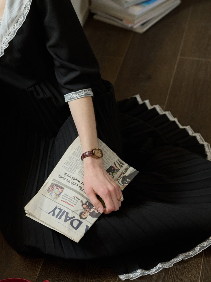 Close-up of a pleated black retro dress with lace hem, styled with a newspaper and classic wristwatch for an elegant and nostalgic outfit detail.