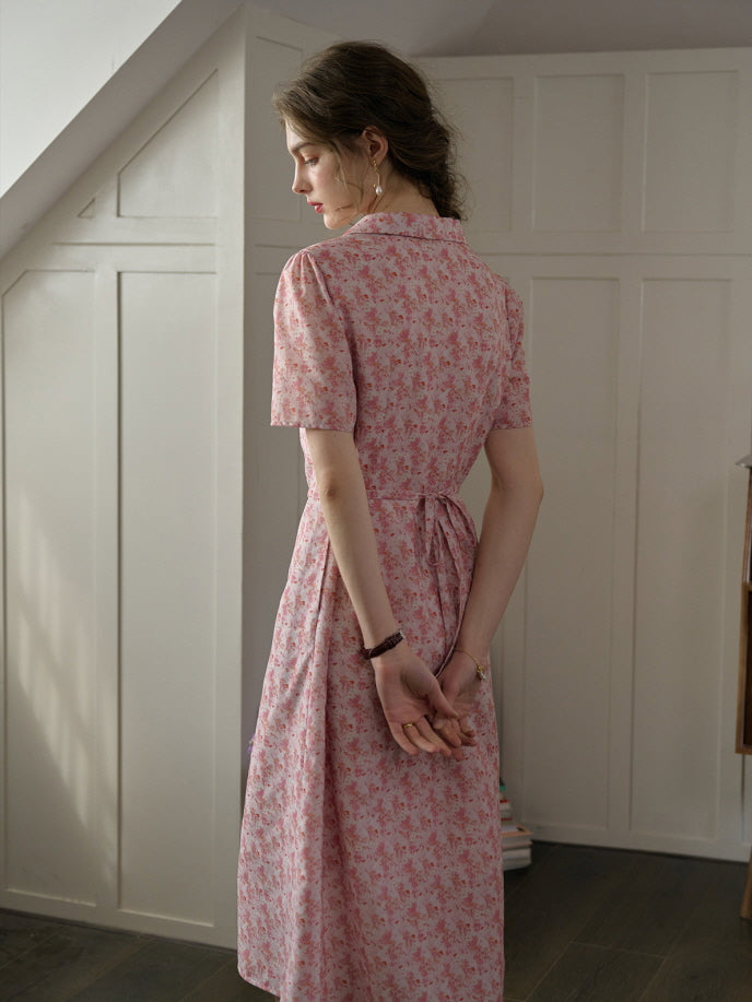 A woman wearing a pink floral cottagecore dress with a tied waist, captured from behind in a soft and casual indoor setting.