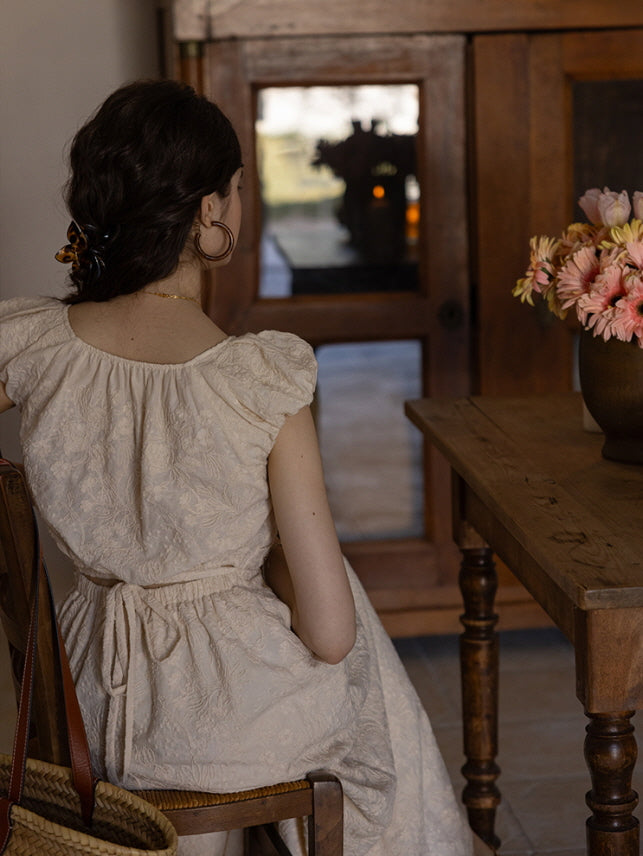 Back view of a woman sitting in a cream-colored textured midi dress with a tied waist and puff sleeves, her hair styled with a tortoise shell clip. A vintage wooden cabinet and a vase of flowers enhance the cottagecore ambiance.