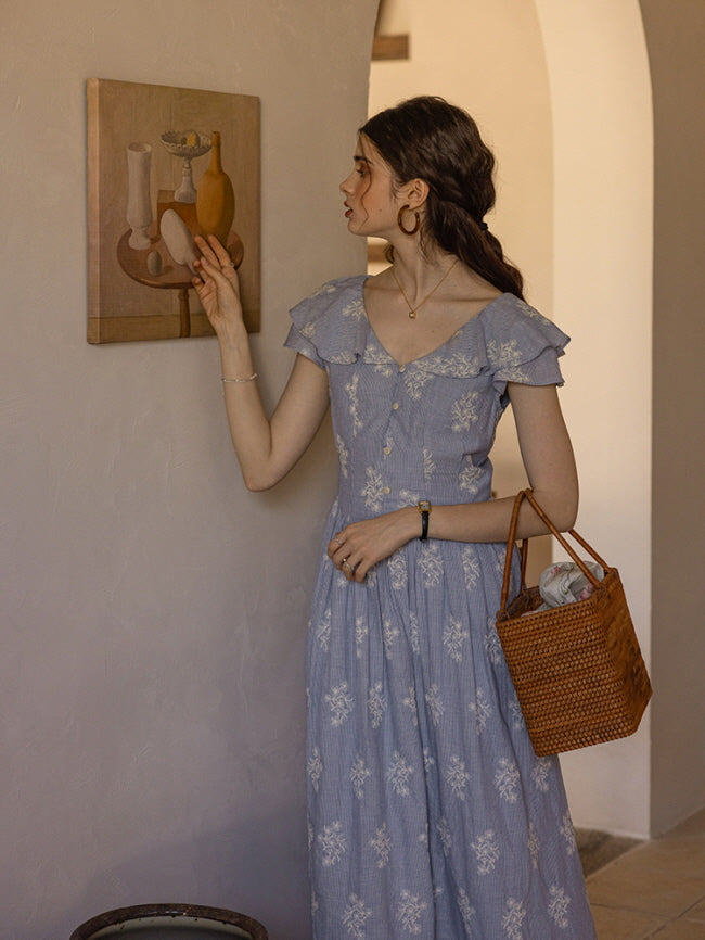 A young woman with soft, wavy hair gently touches a still life painting, her expression calm and reflective. She wears a light blue floral cotton dress with ruffled sleeves and holds a woven rattan bag, capturing a serene, cottagecore ambiance in a vintage-inspired hallway.