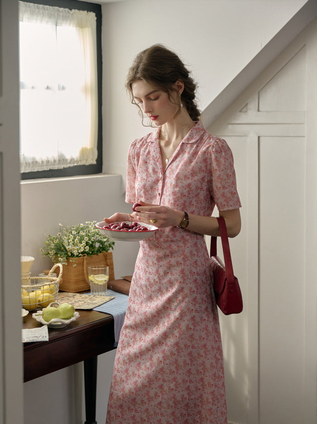 A soft cottagecore outfit featuring a pink floral dress, styled casually with a red shoulder bag as the woman enjoys cherries in a sunlit country-style kitchen.