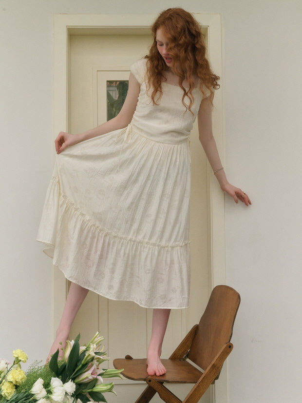 A young woman with red curly hair smiles while standing barefoot on a wooden chair, lifting the skirt of her flowing cream dress adorned with delicate ruffles.