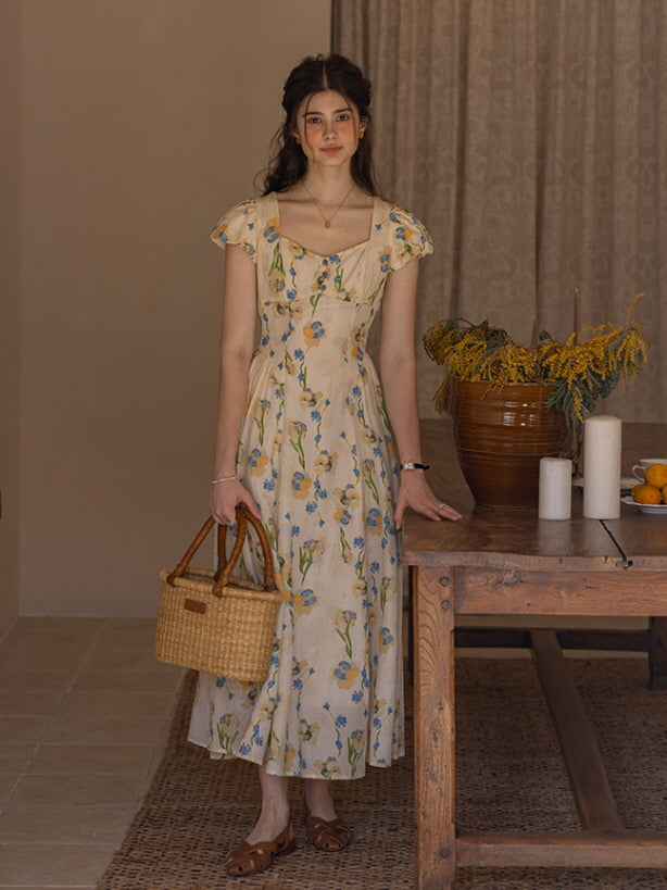 A young woman stands gracefully beside a rustic wooden table, holding a woven basket bag. She wears a cream floral dress adorned with blue and yellow blossoms, exuding a soft, vintage-inspired charm in a cozy, cottagecore setting with dried flowers and candle accents.