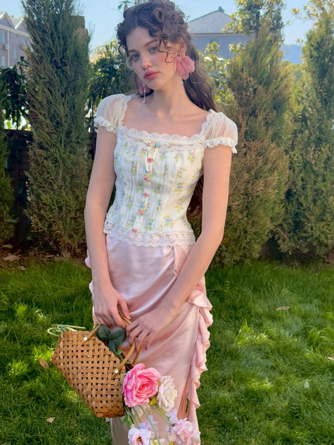 A model stands in a garden holding a woven basket adorned with roses, wearing a floral corset top with lace detailing and sheer puff sleeves, paired with a pink ruched satin skirt for a vintage-inspired look.