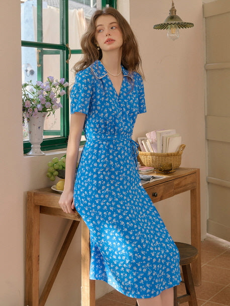 Woman in a royal blue floral dress with a pearl-trimmed collar, sitting by a wooden desk near a green window. Sunlit vintage interior setting with flowers and fruits adds a soft, romantic feel.