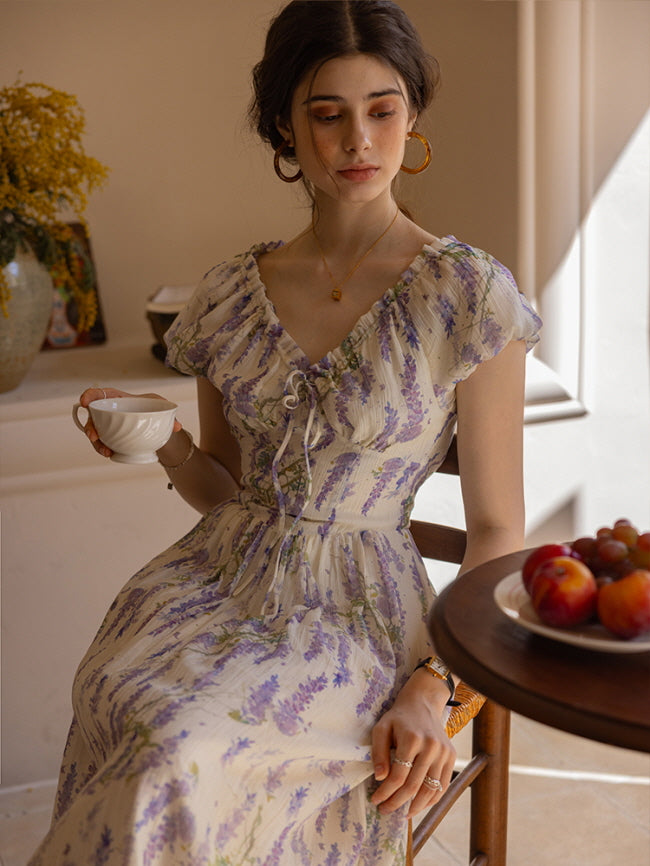 Young woman seated by a window, holding a teacup while wearing a lavender floral dress with lace-up bodice details, creating a soft, cottagecore-inspired atmosphere.