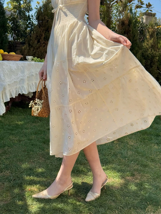 Woman in a romantic fairytale-style cream eyelet dress lifting the hem slightly while standing on a grassy garden lawn, surrounded by a lace-covered table and flowers – cottagecore, princesscore aesthetic.
