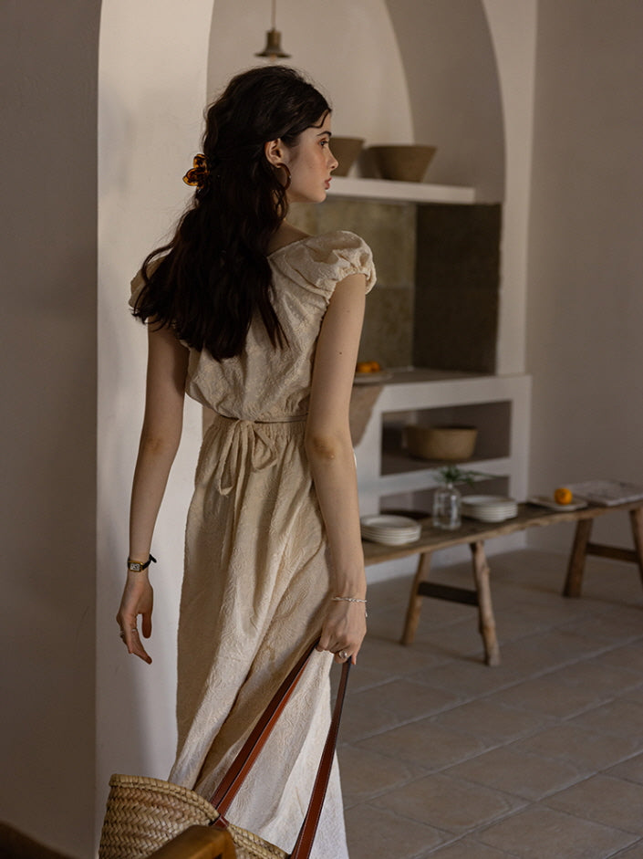 Back view of a woman wearing a cream-colored textured maxi dress with a tied waist, carrying a woven tote bag while walking through a softly lit, minimalist interior space. The atmosphere reflects a calm, vintage-inspired aesthetic.
