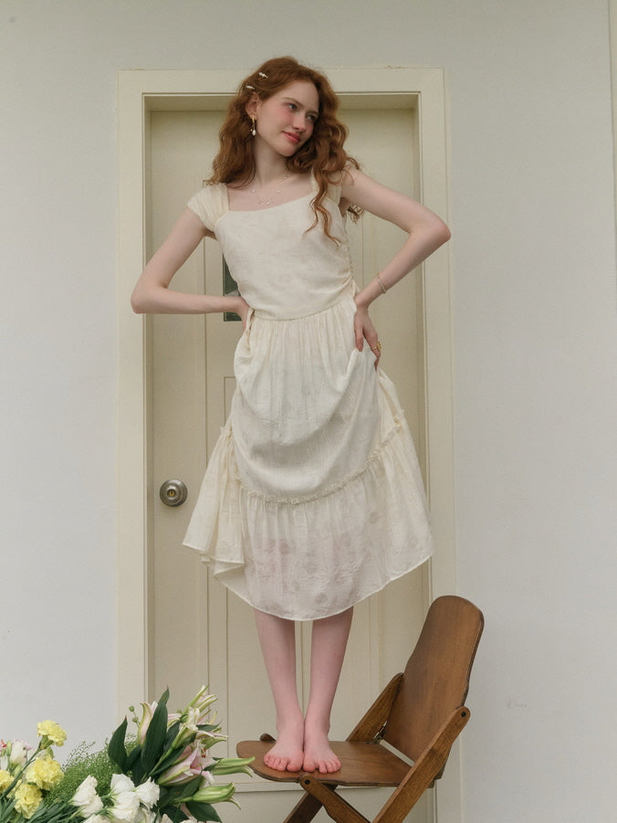 A young woman with red curly hair stands barefoot on a wooden chair, wearing a cream-colored fairy-inspired dress with ruffle details, posing playfully with her hands on her waist.