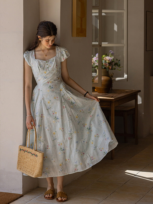 Romantic cottagecore floral dress with cap sleeves and button-down front, styled with a woven straw bag and sandals, creating a soft, fairytale-inspired atmosphere in a rustic, light-filled room.