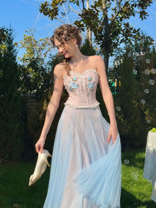 Woman smiling in a pastel floral corset dress with light blue tulle skirt, holding heels in one hand and walking barefoot through a garden with bubbles – whimsical fairytale look