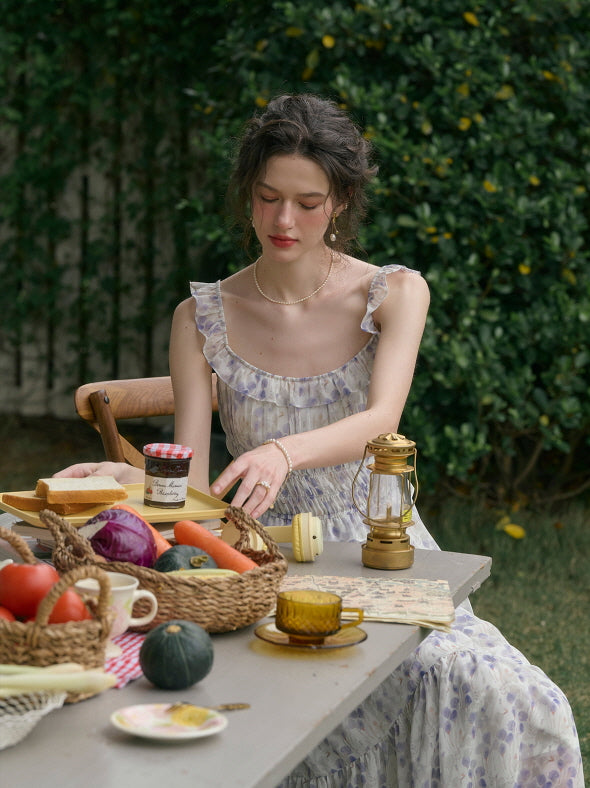 A woman wearing a lavender floral cottagecore dress sits at an outdoor table decorated with rustic picnic food and vintage props.