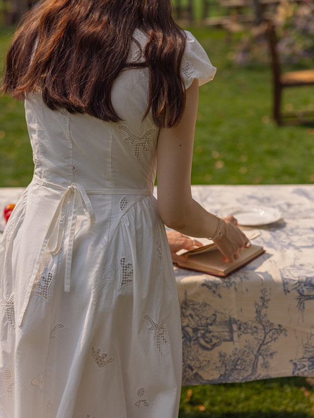 Back view of a woman wearing a white embroidered dress with a lace-up back detail, leaning over a table set with a book and tableware, creating a serene, summer picnic atmosphere.
