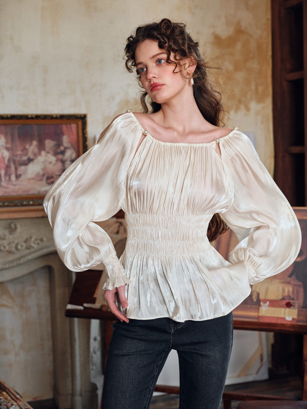 A woman poses confidently in a romantic ivory puff-sleeve blouse with pearl-embellished cutout shoulders, paired with black jeans, in a vintage artist's studio. Her wavy hair and soft makeup enhance the fairytale-inspired, feminine aesthetic.