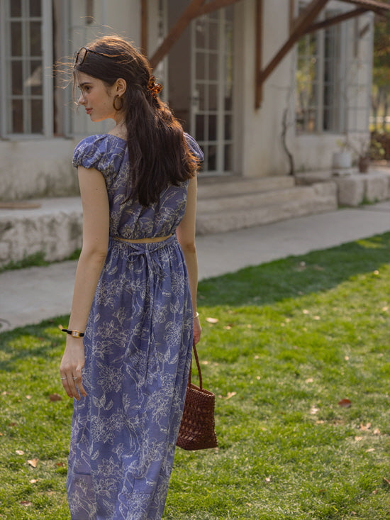 Back view of a blue floral dress with a tie-back detail, as the woman carries a woven bag and walks through a sunlit garden, exuding a romantic cottagecore vibe.