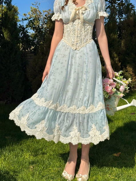 A woman wearing a light blue vintage-style dress with ivory lace trim and a corset bodice, holding a bouquet of pink flowers, with the hem of the dress flowing gently as she stands on grass.