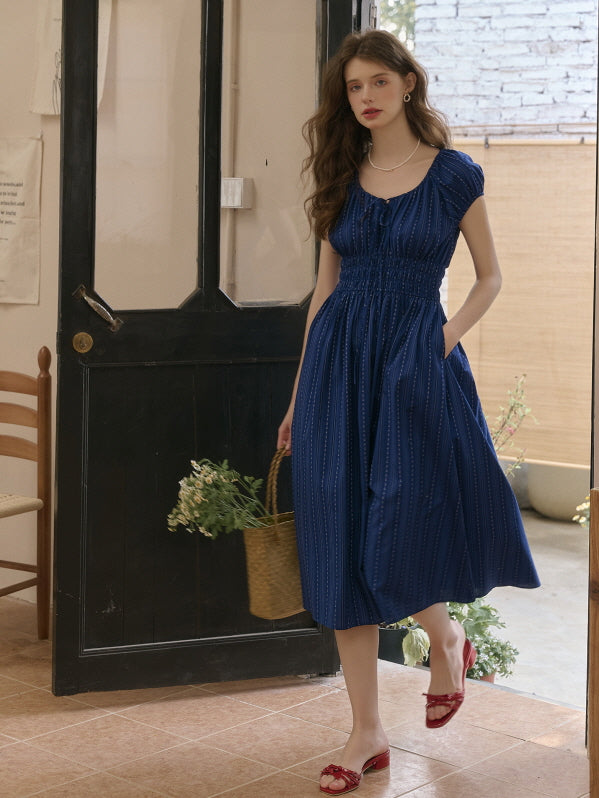 Model wearing a gathered navy midi dress with puff sleeves, holding a straw bag with flowers while walking past a black wooden door. Cozy countryside aesthetic with graceful movement.
