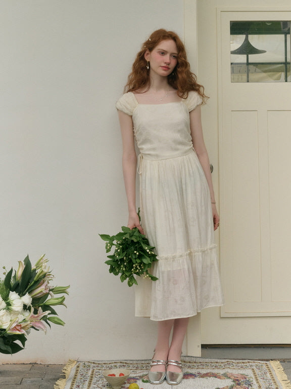 A young woman with long red hair stands barefoot on a patterned rug, wearing a romantic ivory dress with puff sleeves and a ruched waist detail, holding a bouquet of greenery in one hand. She poses gracefully in front of a white door, surrounded by soft floral decorations.