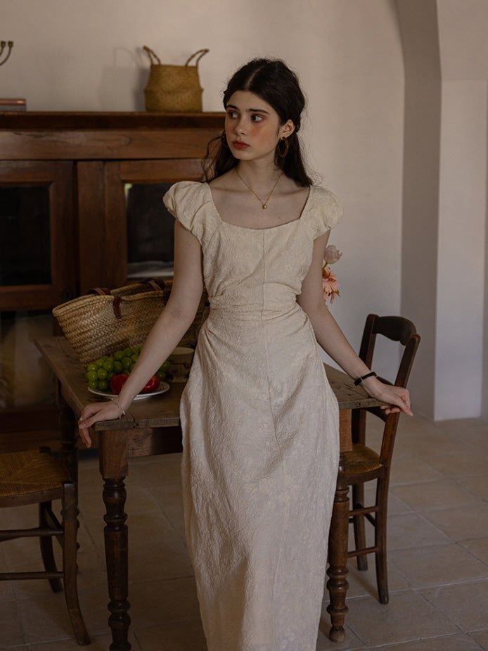 Woman in a cream-colored textured maxi dress with puff sleeves and a square neckline, leaning against a wooden table with a woven basket and a plate of grapes. The setting exudes a rustic, cottagecore ambiance.