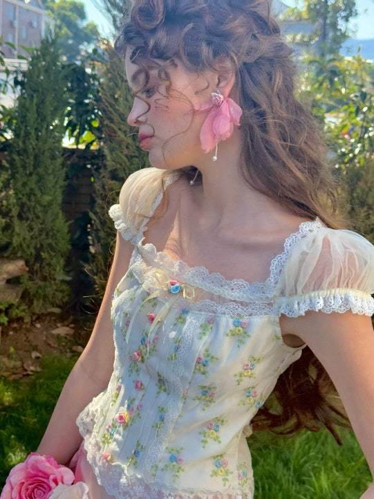 A close-up of a floral corset top with lace trim and sheer puff sleeves, featuring delicate pastel floral embroidery and ribbon details, styled with a pink ruched satin skirt for a romantic, fairytale look.