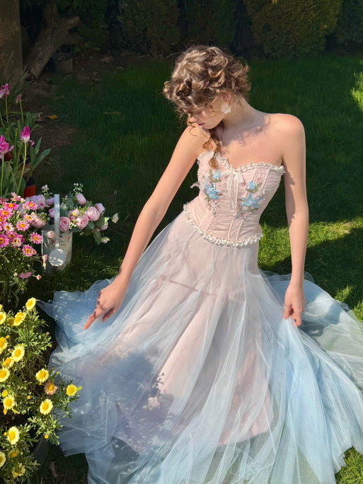 Romantic woman in a pastel blue tulle corset dress with floral embroidery, sitting in a garden surrounded by flowers – fairytale-inspired outfit with soft elegant silhouette