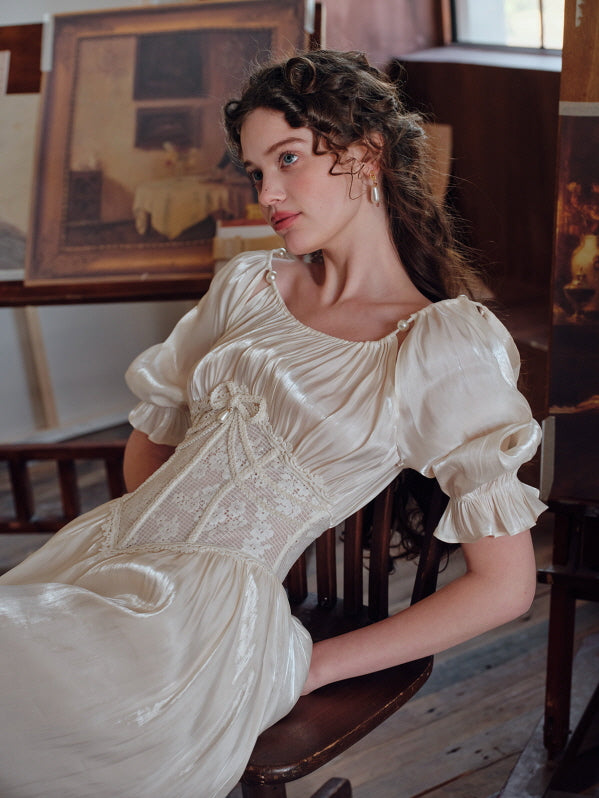 A woman leaning back on a wooden chair, wearing a pearl-accented ivory dress with puff sleeves and an embroidered lace corset, styled in a softly lit vintage art studio.